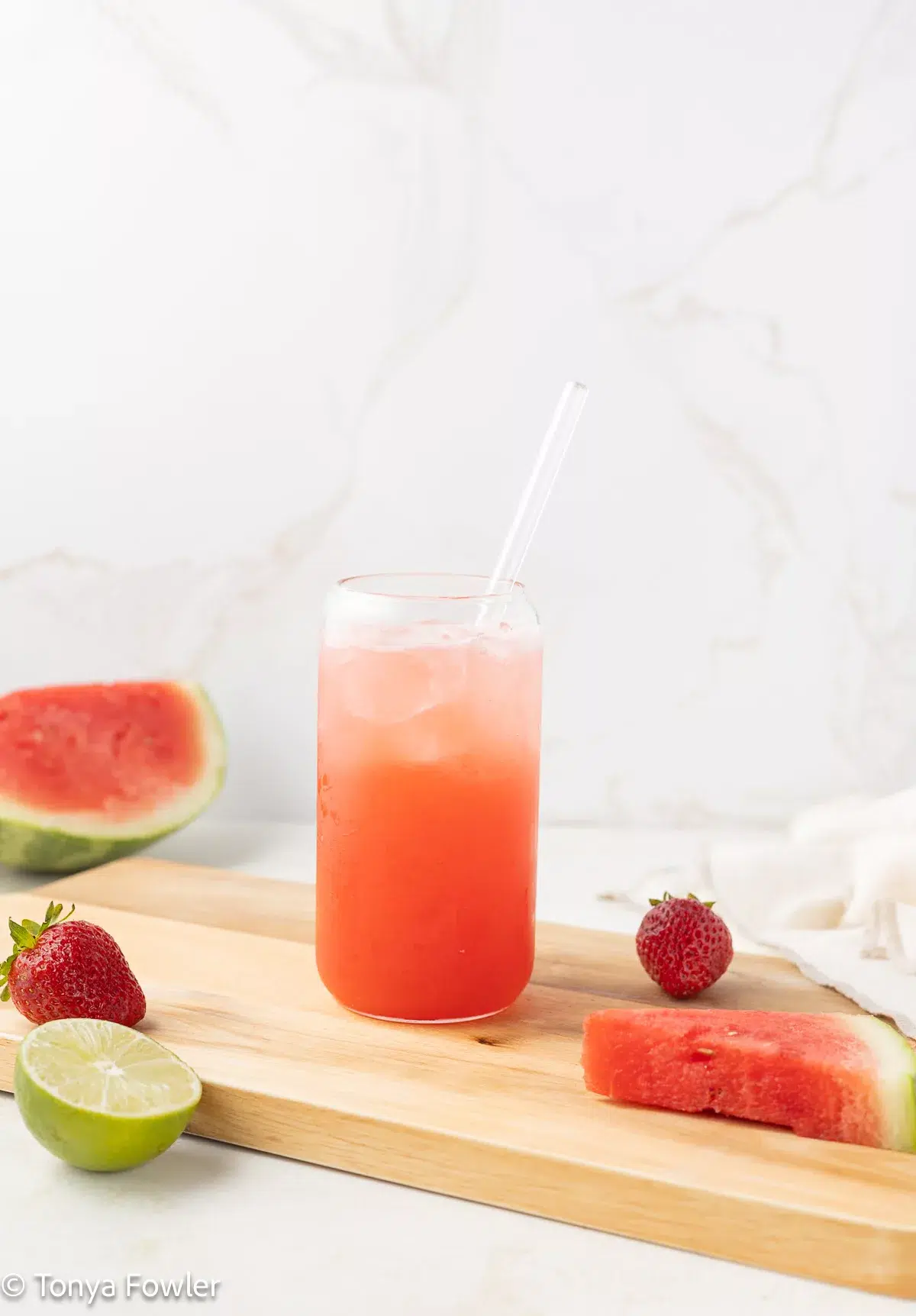 Straight on image of a watermelon strawberry mocktail in a glass sitting on a cutting board with fresh fruit all around.