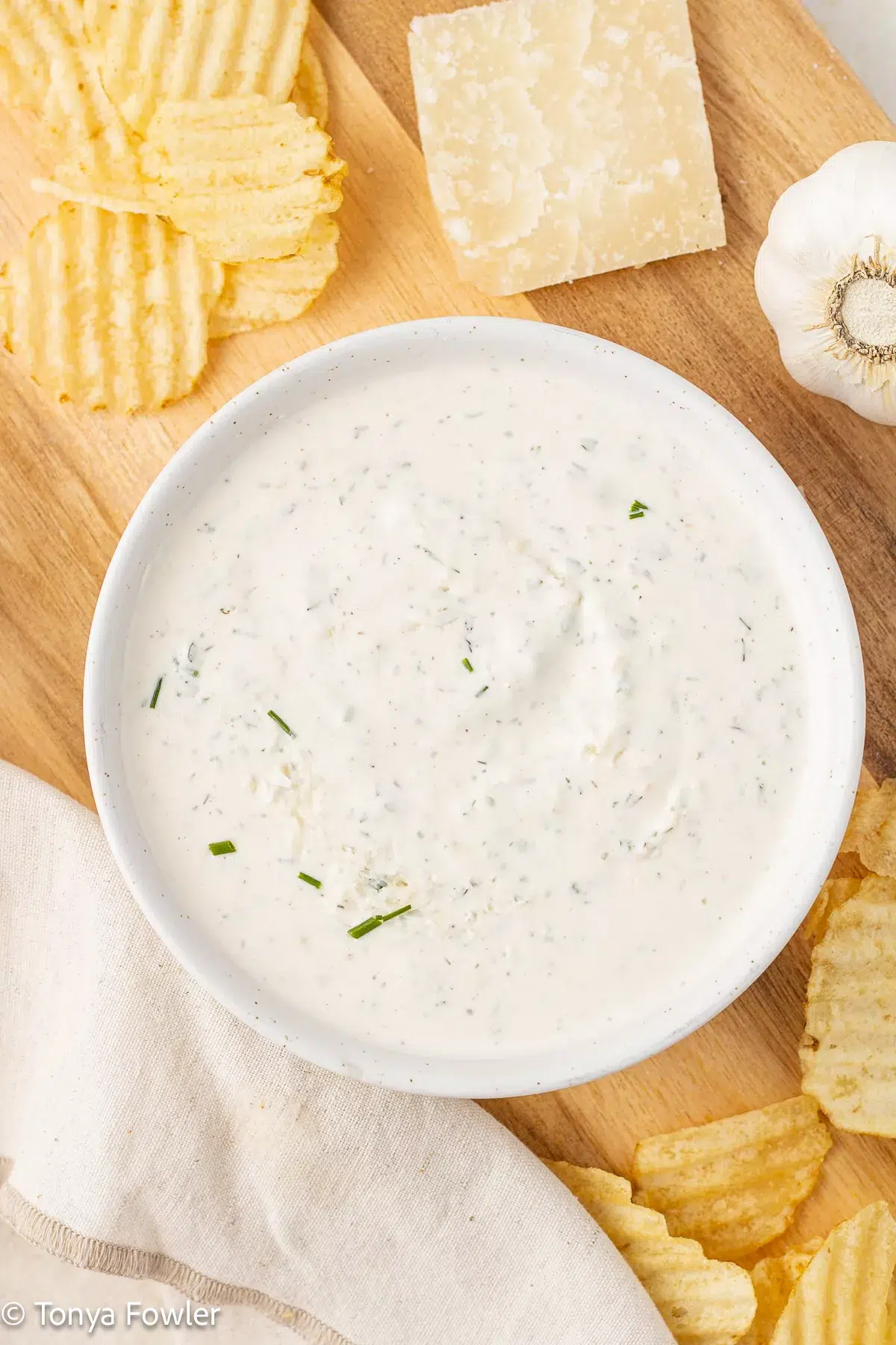 Overhead image of ranch dip in a bowl surrounded by potato chips, parmesan cheese and garlic.