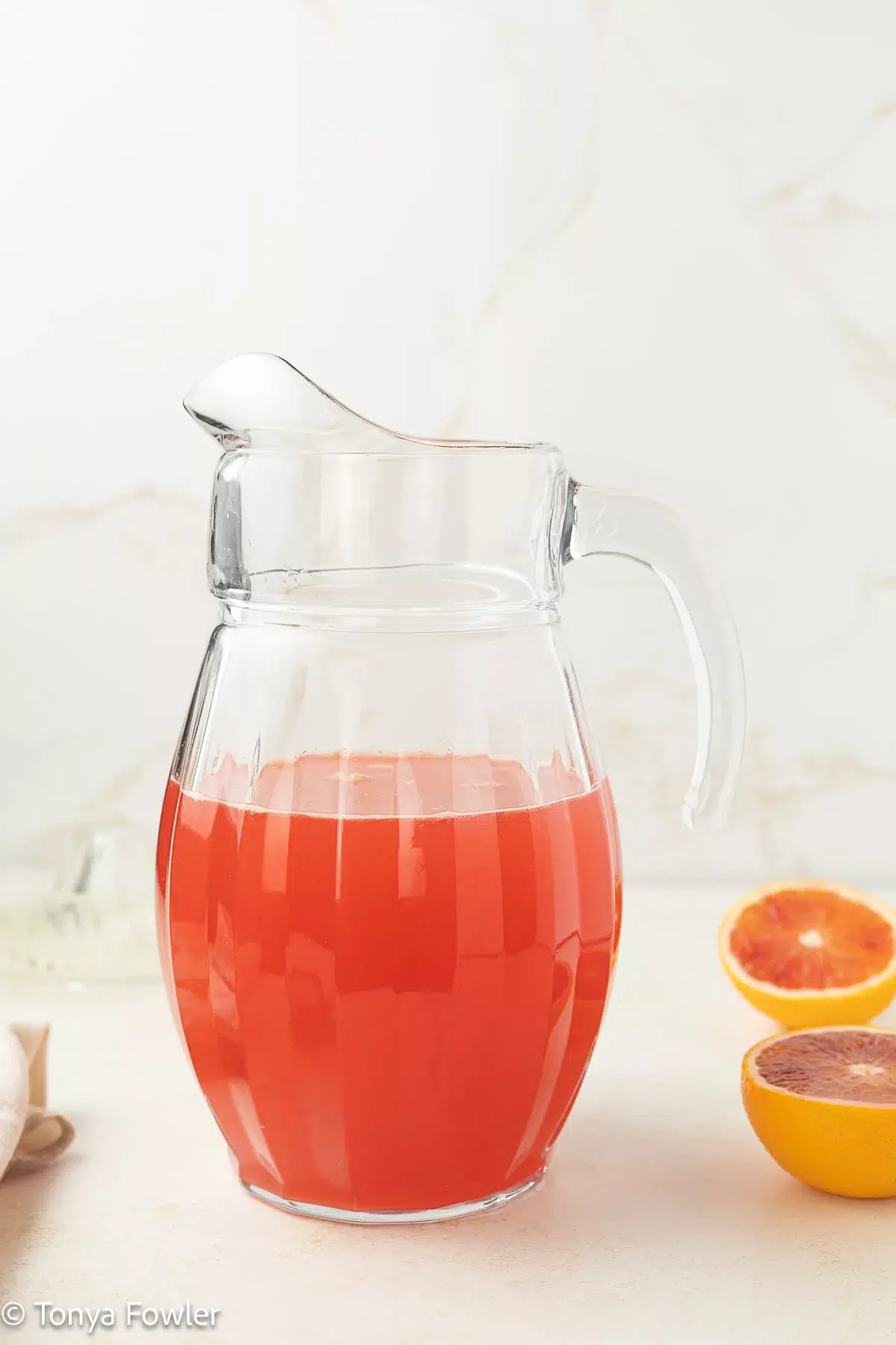 Orange lemonade in a pitcher with sliced oranges in the background.