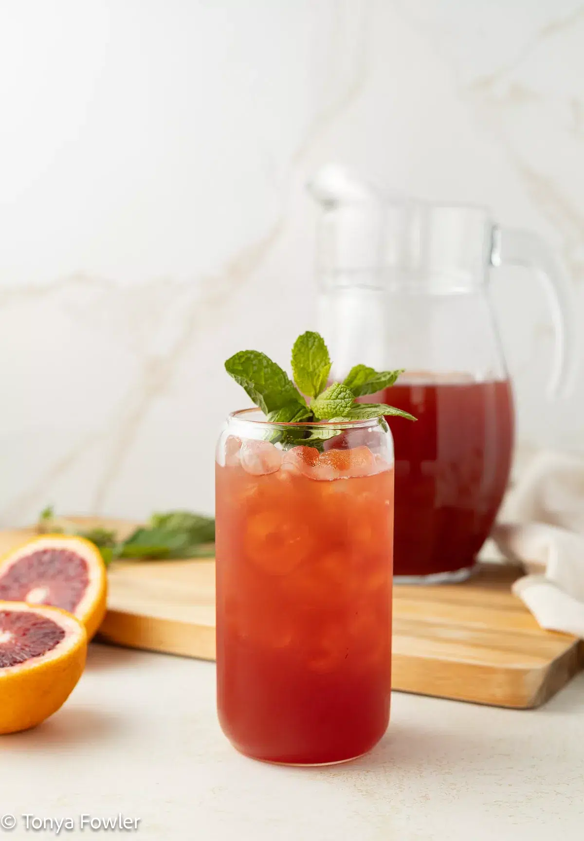 Straight on image of blood orange iced tea in a glass with mint and fresh fruit on the counter.
